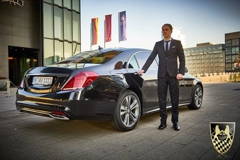 A professional chauffeur dressed in formal attire stands beside a sleek black Mercedes-Benz S-Class sedan, parked in front of a grand building in Munich, symbolizing luxury and exceptional service for airport transfers and business trips. The scene captures the essence of a stress-free travel experience, ideal for those seeking a private driver in Munich.